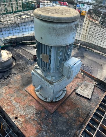 An industrial electric motor and gearbox, covered in a layer of dirt, are mounted on a corroded and rusty metal platform. The motor's finned casing and terminal box are visible.