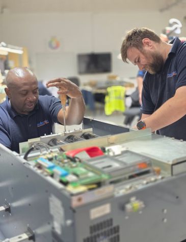 Two Neutronic Technologies engineers are working together to repair a large electronic unit. One engineer is using a screwdriver to tighten a screw on the chassis, while the other is observing and assisting. The unit's internal components, including circuit boards and fans, are visible.
