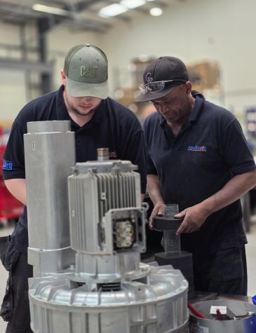 Two Neutronic technicians in dark blue shirts and caps assembling or inspecting a large metallic machine component in an industrial workshop