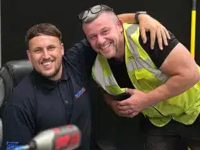 Two Neutronic team members posing in a workshop setting, one seated in a navy polo shirt with company logo, the other standing in a high-visibility vest, with tools visible in the foreground