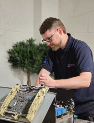 A technician in a navy blue polo shirt repairs a variable speed drive with its internal circuit boards and wiring exposed. The technician’s face is blurred for privacy. A potted plant and textured white wall are visible in the background.