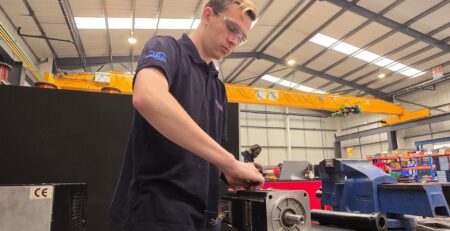 Technician conducting servo motor repair and maintenance in an industrial workshop, surrounded by tools, equipment, and supply components including a blue vise and overhead crane system.