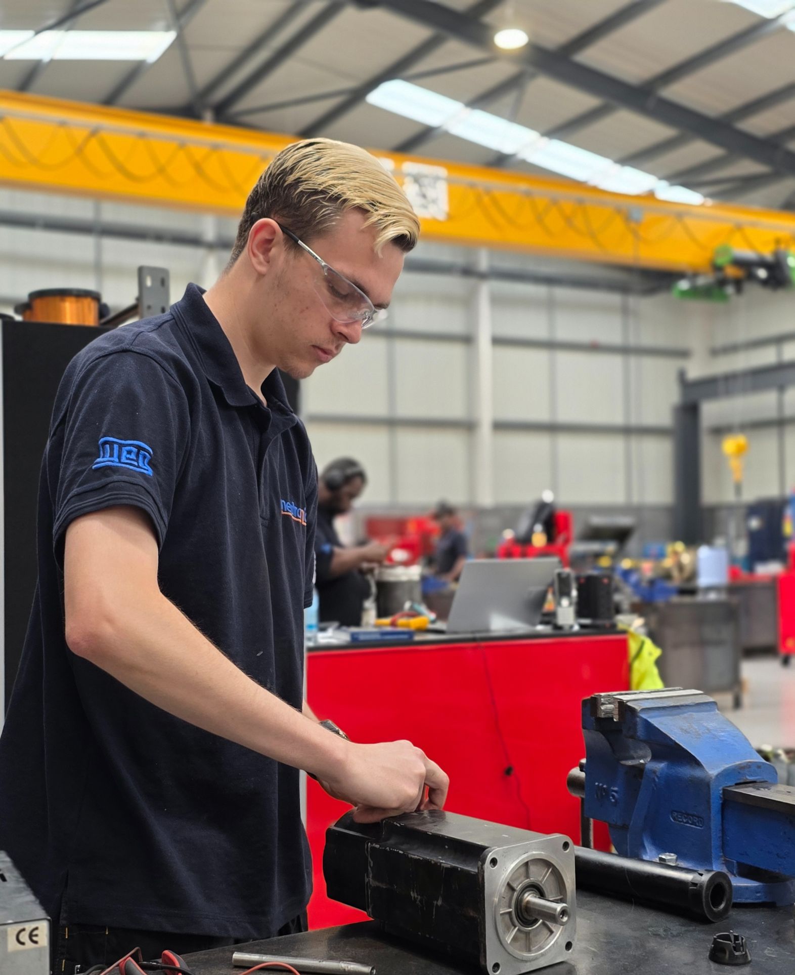Technician performing servo motor repairs in a workshop, wearing a dark blue uniform. The workspace includes tools, a blue vise, and an overhead crane system, emphasizing a professional repair environment.